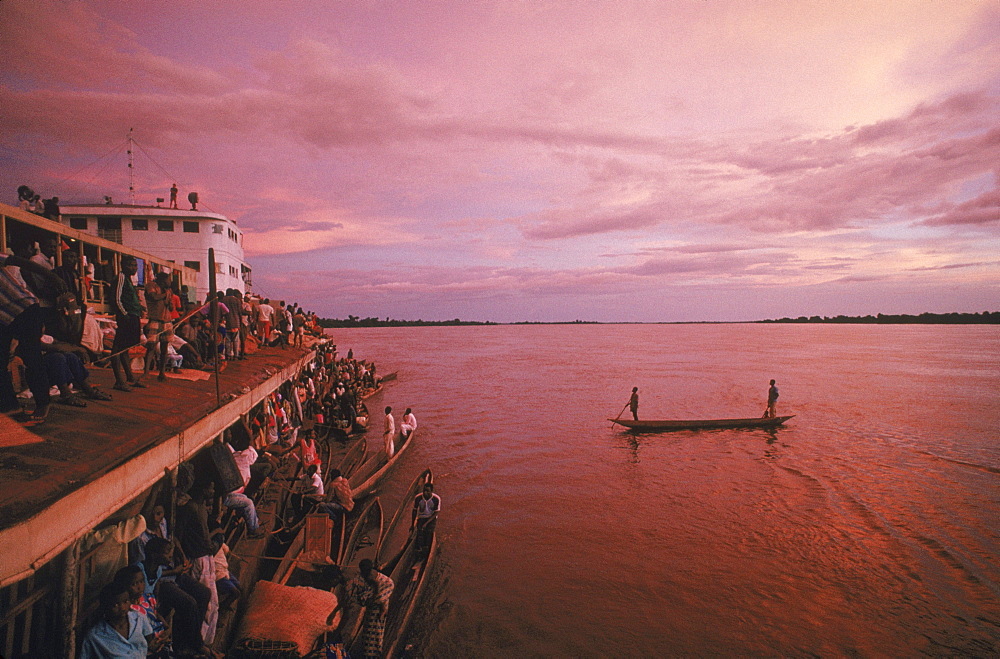 Men in a canoe paddle to join with others tied up the the side of barges pushed up the Congo River by the Col. Ebeya. The canoes bring fish, meat, insects, and produce to the boat, which the fishermen trade for manufactured goods.