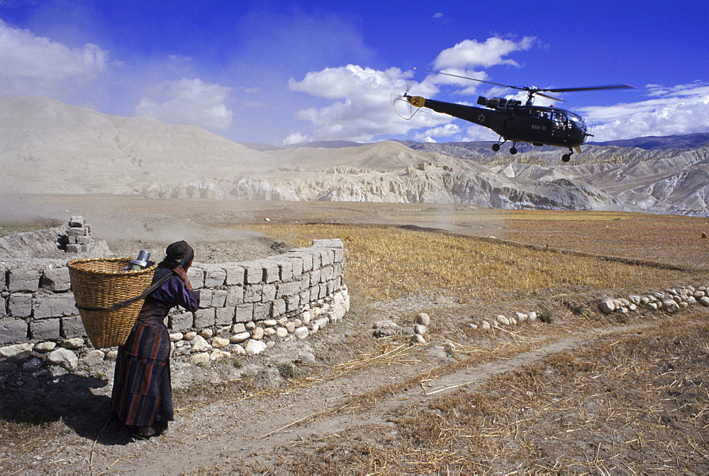 Woman with grain basket watches as a helicopter takes tourists out of the remote Kingdom of Mustang, Nepal.