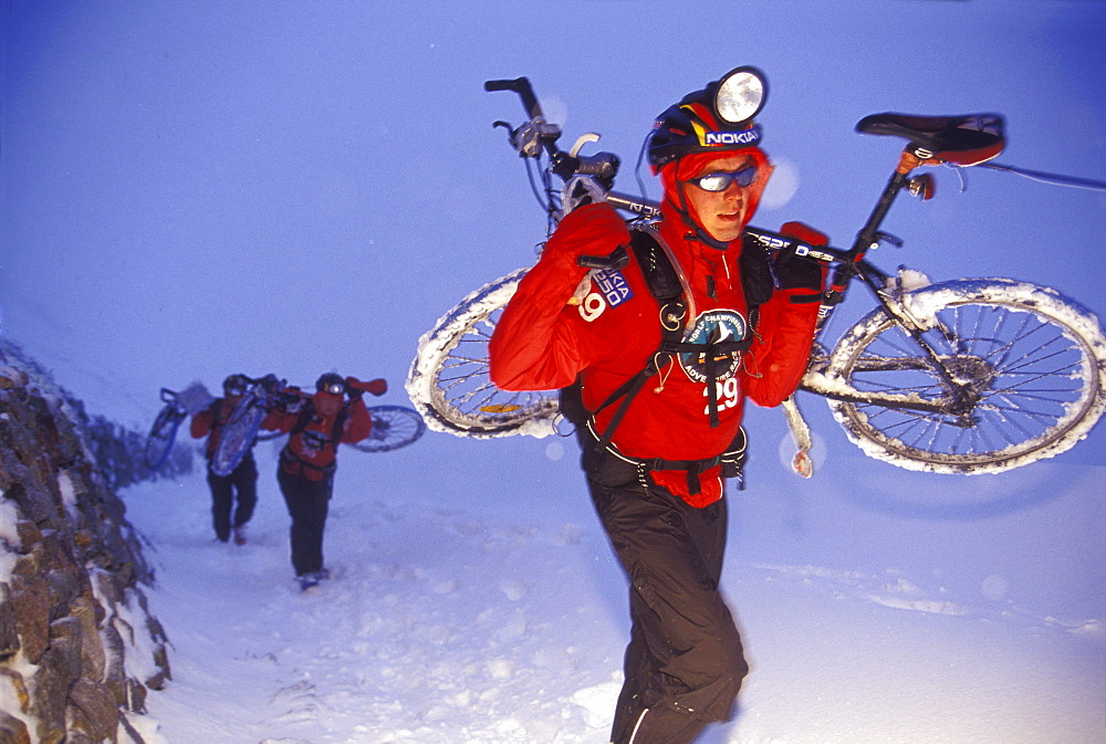 A man carries icy mountain bike through a snowstorm during the 2001 Adventure Racing World Championships in Switzerland, two other team members in the background