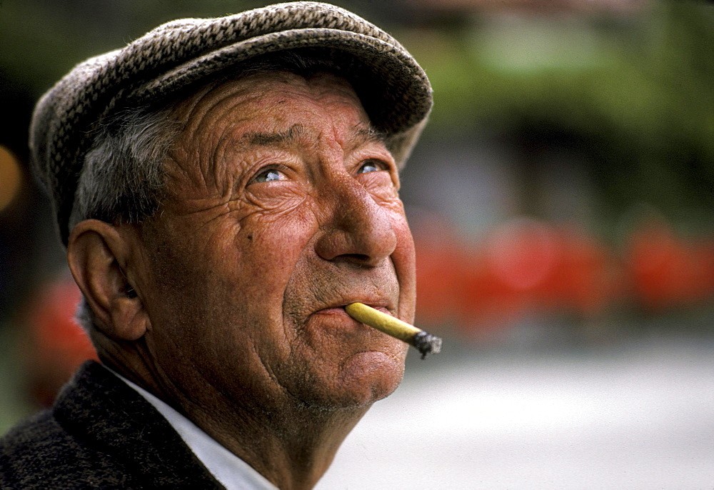 An older man holds a cigarette between with teeth and smiles.