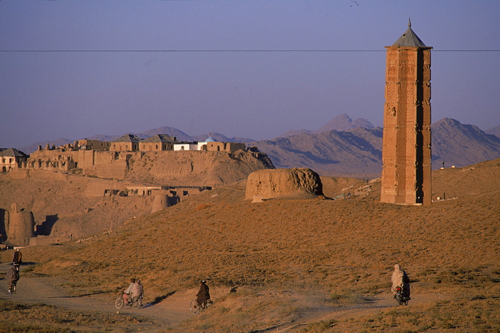 Men on bikes ride past an ancient  brick minaret  toward the old walls and citadel of the town of Ghazni, Afghanistan, October 1, 2002. Made of brick decorated with Kufic and Naksh Script and floral motifs, the  minaret dates back to  the early 12th century and was built by Sultan Masud III of the Ghaznavid Dynasty, who ruled over an empire encompassing much of Afghanistan, Northern India, Persia and Central Asia. The minaret was once three times as tall as its current 70 feet, and is thought to have been part of a large mosque complex. Now an  important truck stop on the road to Kandahar, Ghazni, located on the Lora River at the elevation of 2,225 meters, is the capital of Ghazni province with a population of 35,900, and is a  market for sheep, wool, camel hair cloth, corn, and fruit.