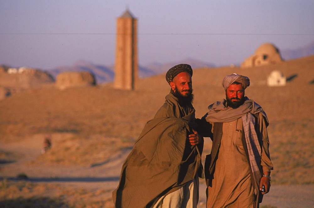 Afghan men pause on the road with an ancient  brick minaret  and many shrines in the background, outside of Ghazni, Afghanistan, October 1, 2002. Made of brick decorated with Kufic and Naksh Script and floral motifs, the  minaret dates back to  the early 12th century and was built by Sultan Masud III of the Ghaznavid Dynasty, who ruled over an empire encompassing much of Afghanistan, Northern India, Persia and Central Asia. The minaret was once three times as tall as its current 70 feet, and is thought to have been part of a large mosque complex. Now an  important truck stop on the road to Kandahar, Ghazni, located on the Lora River at the elevation of 2,225 meters, is the capital of Ghazni province with a population of 35,900, and is a  market for sheep, wool, camel hair cloth, corn, and fruit, and continues to be a haven for  Taliban insurgents.