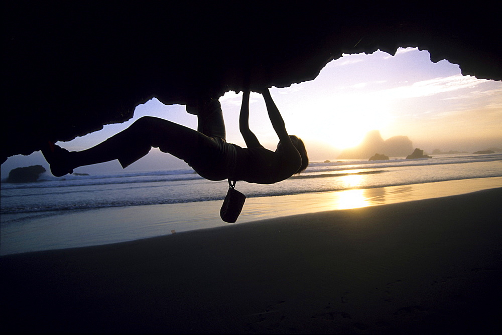 A woman boulderer hangs above the beach at sunset at Houda Pt. in Northern California