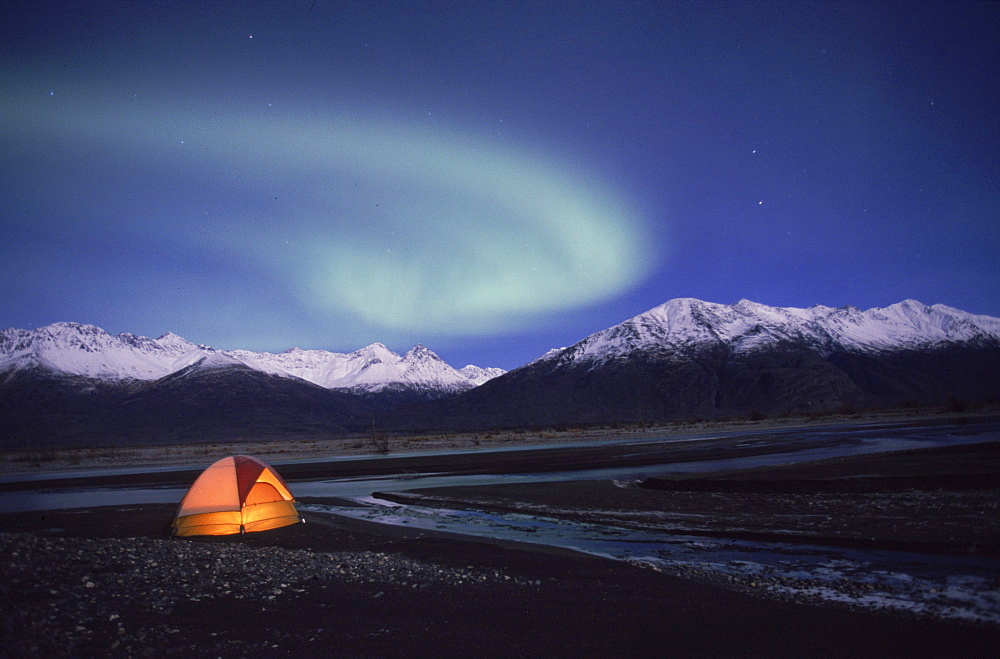 The aurora borealis (northern lights) glows above a tent in the Knik River Valley. The Chugach Mountains are in the background