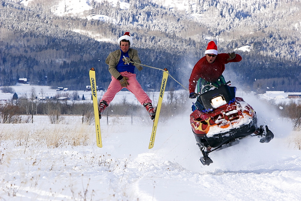 An excited man on a snowmobile gets some air while towing another man in pink snow pants on skis.