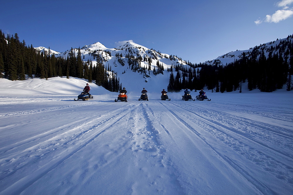 A line of six people on snowmobiles head toward the camera with Owlshead Mountain looming behind them.