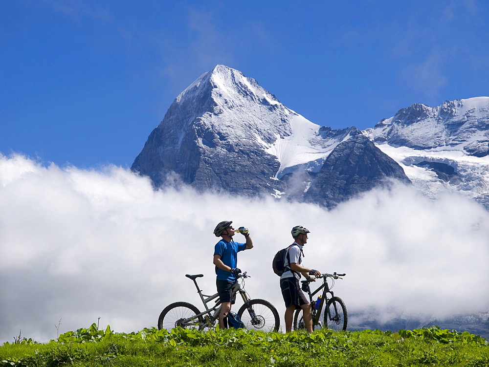 Two mountain bikers are resting and drinking near the village of Murren, during a long bike ride in the Bernese Oberland. In the background the famous Eiger mountain.