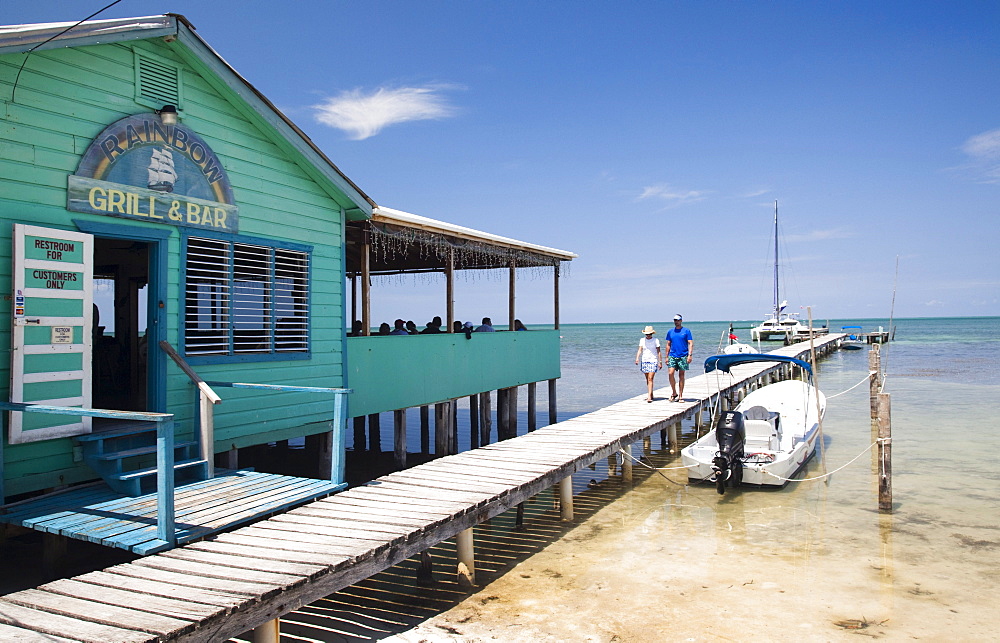 A couple walk down a dock near a ocean side restaurant.