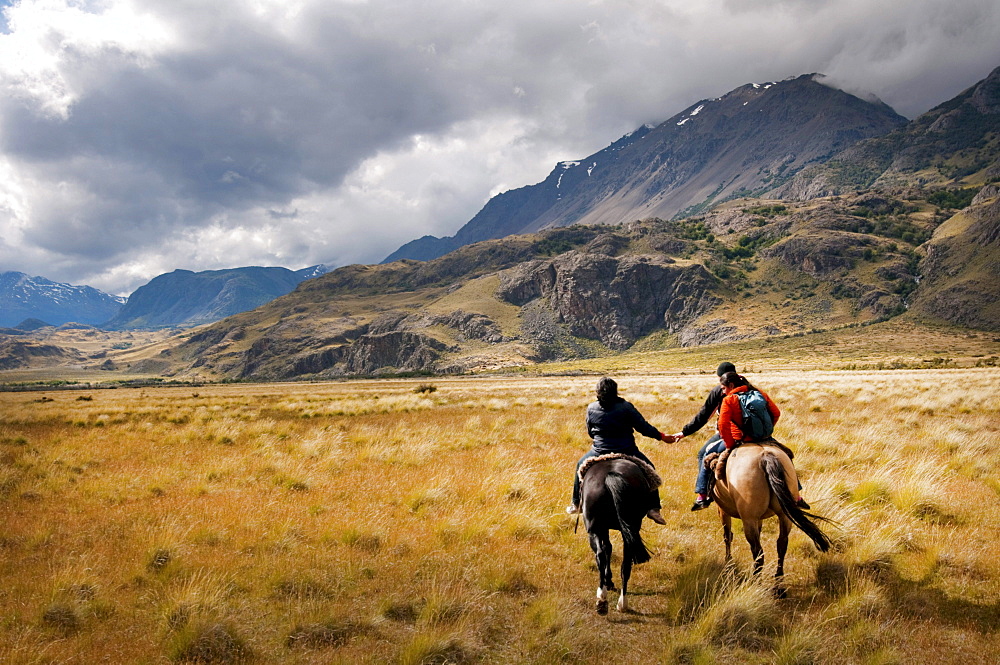 A monther and dayghter hold hands on horse back as they head off to herd sheep on Estancia Chacabuco, Patagonia.