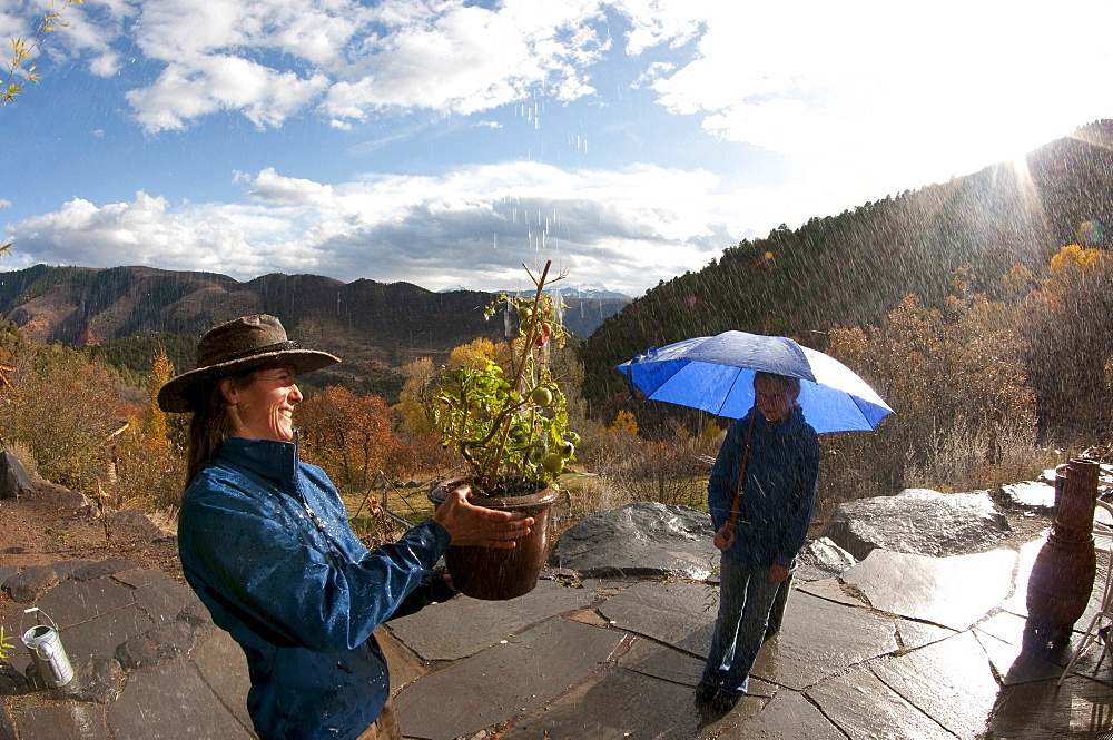 A woman smiles as she waters a tomato plant with rain water with an autumn view of the Colorado mountains. Her daughter stands under an umbrella with the sun shining behind her.