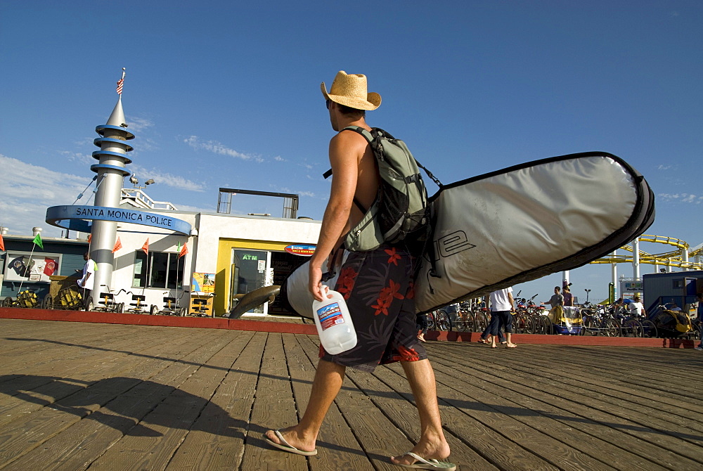 USA/CA/Santa Monica/Pier/September 2007:Santa Monica Pier - a landmark in the USA. A surfer (with board, hat and water bottle) walks down the Pier having left the beach front on a warm late summer's afternoon. The beach front Police Station on the Pier adjacent to the carnival rides. The Pier dates from 1909 and still draws in the crowds. It's a nonstop sideshow of activity with street performers, amusement arcade, aquarium, rollercoaster, ferris wheel and the 1922 Looff Hippodrome Carousel (featured in The Sting). It leaves you wondering how all this fits on just one pier. Glorious views out over the beach front and coastline to be had, especially from the ferris wheel.