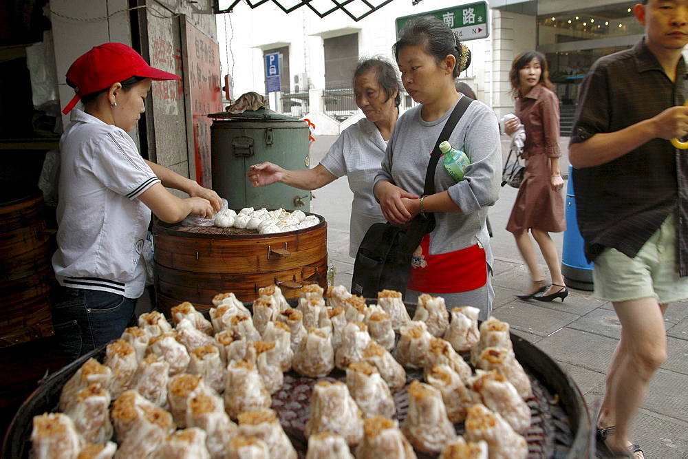 Customers queue up to buy dim-sum style snack foods at a street corner food kitchen near Beijing Dong Lu, central Shanghai, China. In the foreground are shu mei - thin skinned, filled with rice and usually diced pork.