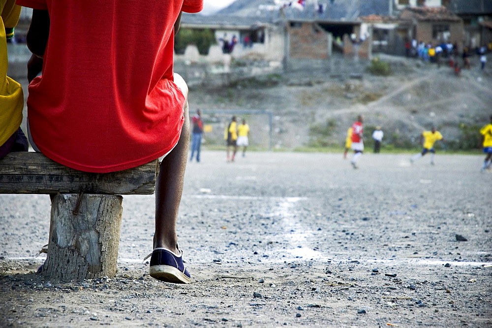 A young man wearing a bright t-shirts sits on a wooden bench watching a soccer game on a rustic dirt field.