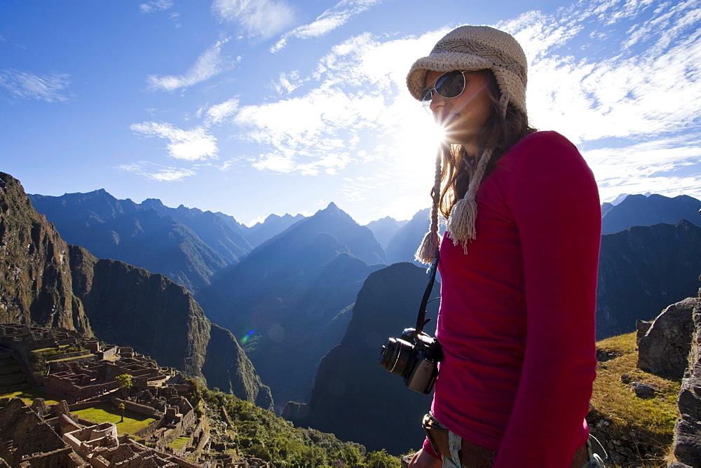 A young girl stands with a camera around her neck overlooking a steep valley, the rising sun, and the ruins of a lost civilization.