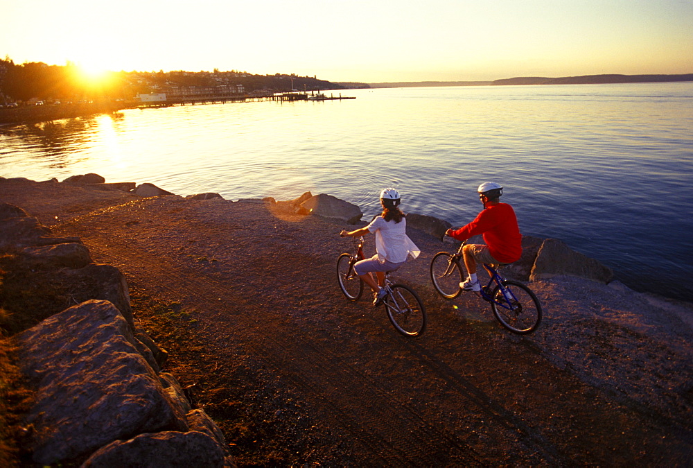 A young couple ride their bikes at sunset along the Puget Sound waterfront in Tacoma, Washington.