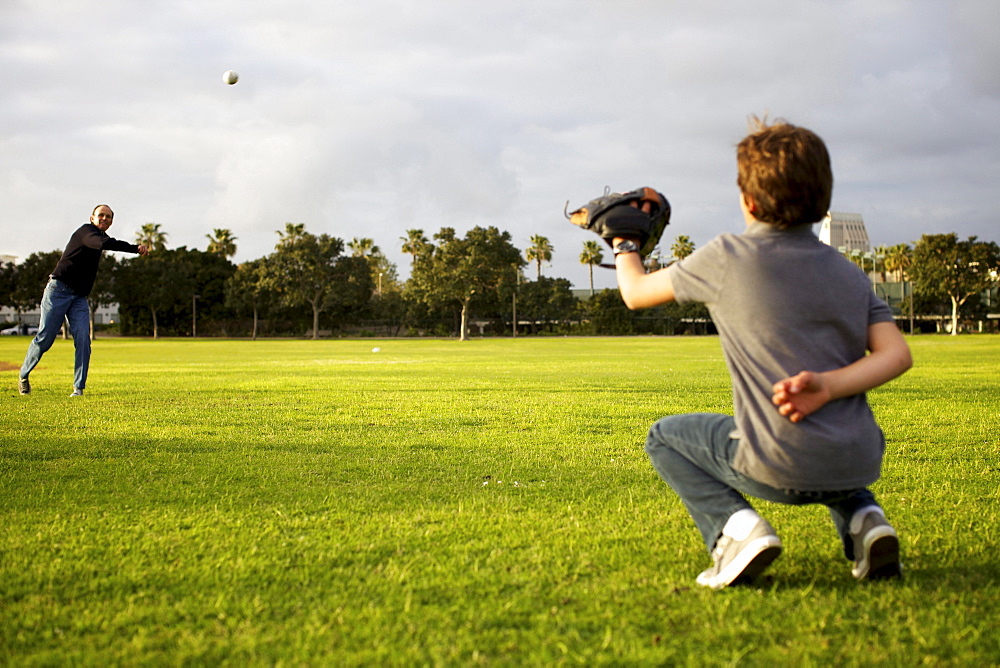 A dad throws a baseball to his kid to catch at the park.