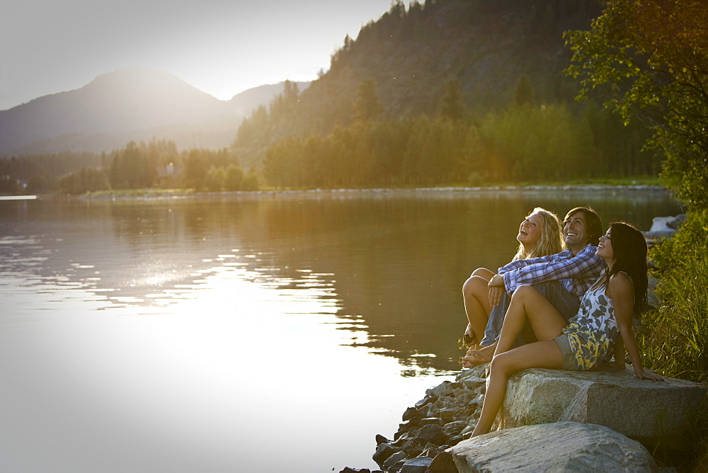 Three young adults laugh while sitting on rocks watching the sunset over the lake.