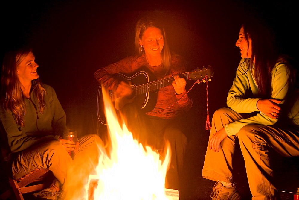 Two women listen, as a third plays the guitar beside a campfire.