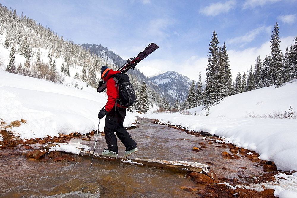 A  man carries his skis across a snowy, mountain stream.