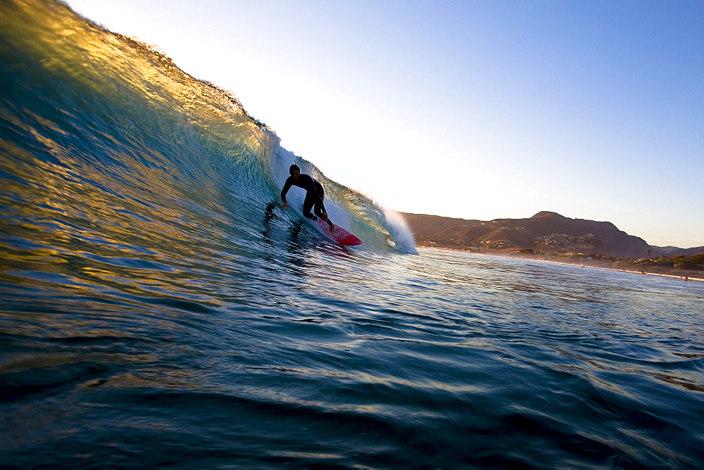 A surfer riding a red surfboard sets up for a barrel while surfing at Zuma Beach in Malibu, California.