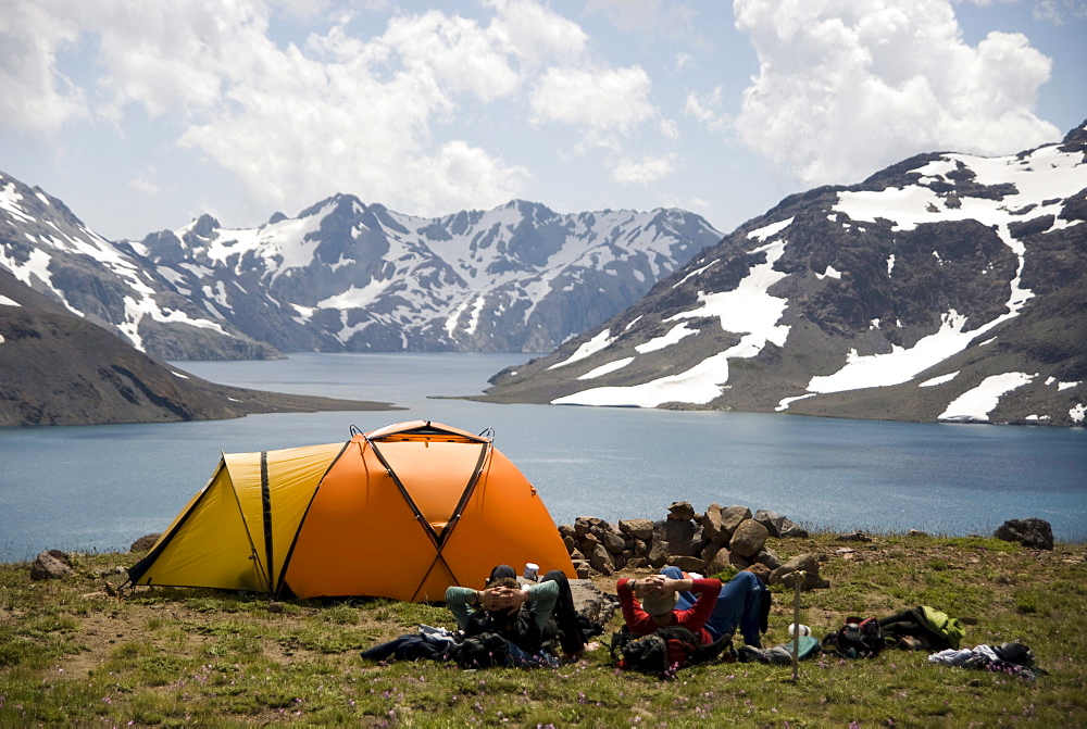 Two men rest next to their orange tent enjoying the dramatic views over a high alpine lake.