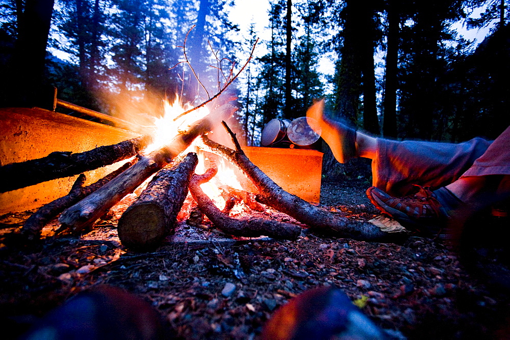 A camper dries her foot next to the flames of their campfire.