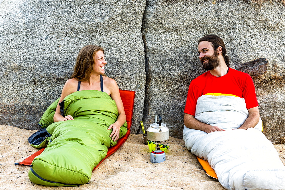 Man and a woman on the beach in sleeping bags laughing with a stove heating water.