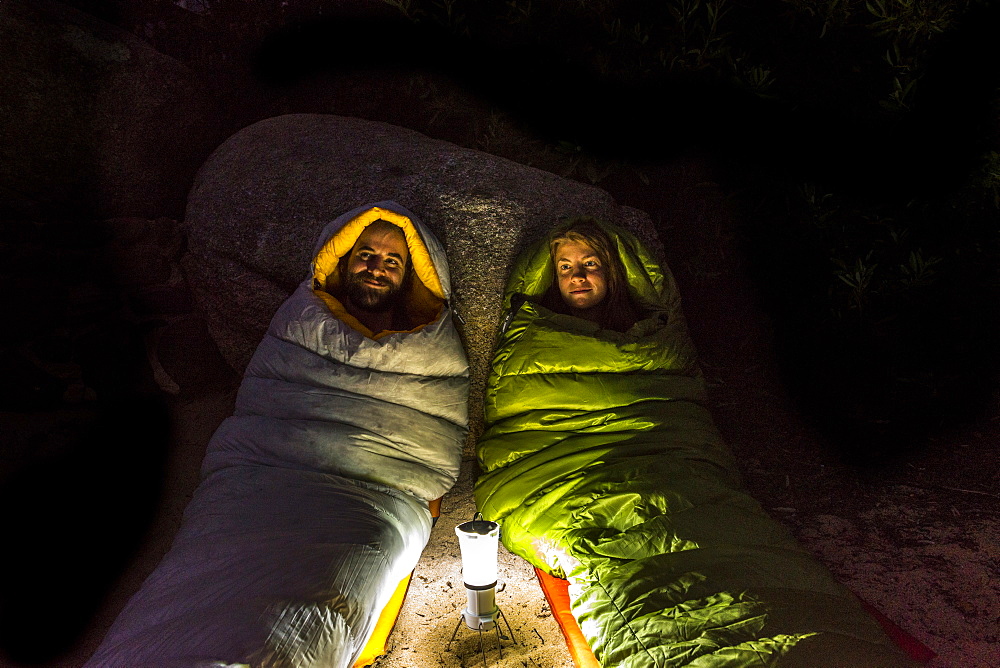 Man and a woman in sleeping bags smiling lit by a lantern at night.