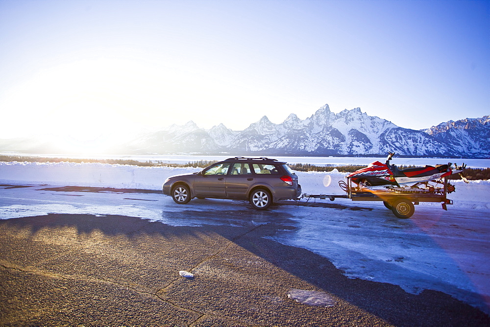 Parked in front of Tetons, a station wagons pulls an older snowmobile on a trailer just before sunset.