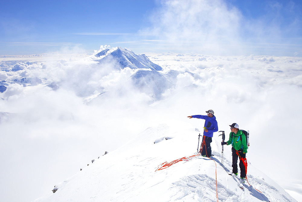 High Mountain Rangers Tom Ditola and David Weber are taking a rest on the West Rib on Mount McKinley, Alaska. Mount Hunter in the background. They are above the clouds in blue sky and the sun is shining.     Mount McKinley, native name Denali, is the highest mountain peak in North America, with a summit elevation of 20,321 feet (6,194 m) above sea level. At some 18,000 feet (5,500 m), the base-to-peak rise is considered the largest of any mountain situated entirely above sea level. Measured by topographic prominence, it is the third most prominent peak after Mount Everest and Aconcagua. Located in the Alaska Range in the interior of the U.S. state of Alaska, McKinley is the centerpiece of Denali National Park and Preserve.    Every climbing season High Mountain Rangers of the Denali National Park Service are called to help climbers in need. If possible the patient is brought down to base camp on foot, only in life threatening conditions a helicopter is called to evacuate the patient to a hospital in Anchorage.