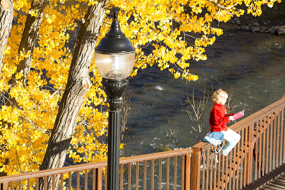 A young girl reading a book while sitting on a bridge over the Animas River on the Animas River Trail in Durango, Colorado.