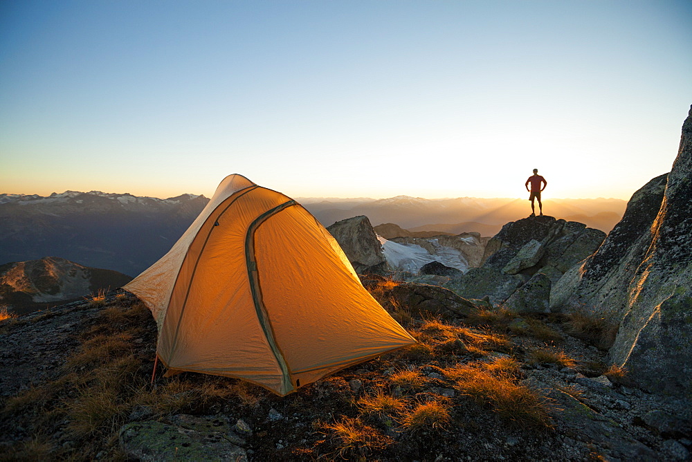A silhouetted hiker stands on a rock bluff while camping on the summit of Saxifrage Peak, Pemberton, BC, Canada.