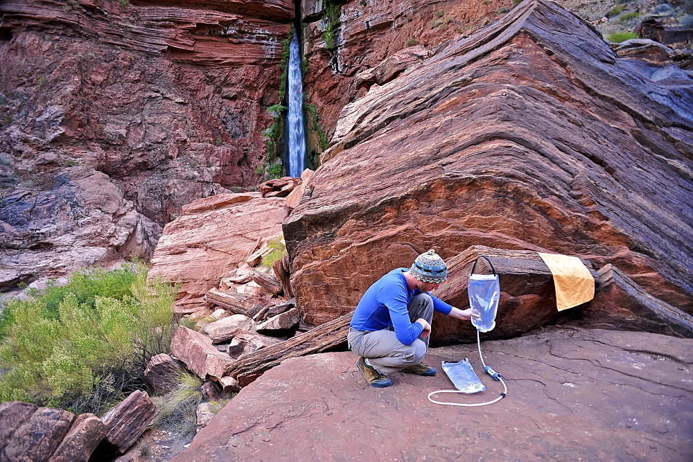 Male hiker filters water on a cliff-pinched patio near Deer Creek Falls in the Grand Canyon outside of Fredonia, Arizona November 2011.  The 21.4-mile loop starts at the Bill Hall trailhead on the North Rim and descends 2000-feet in 2.5-miles through Coconino Sandstone to the level Esplanada then descends further into the lower canyon through a break in the 400-foot-tall Redwall to access Surprise Valley.  Hikers connect Thunder River and Tapeats Creek to a route along the Colorado River and climb out Deer Creek.