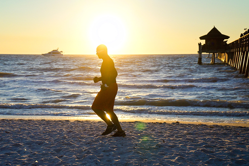 Man jogging on the beach at sunset with a yacht on the horizon and a pier in the background