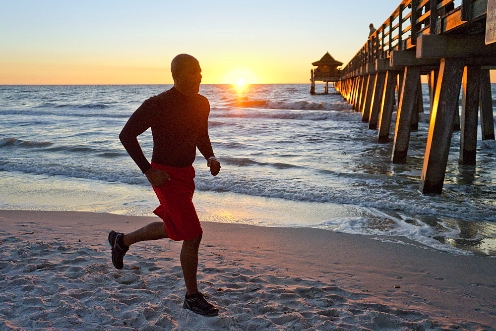 Man running on the beach at sunset with a pier in the background