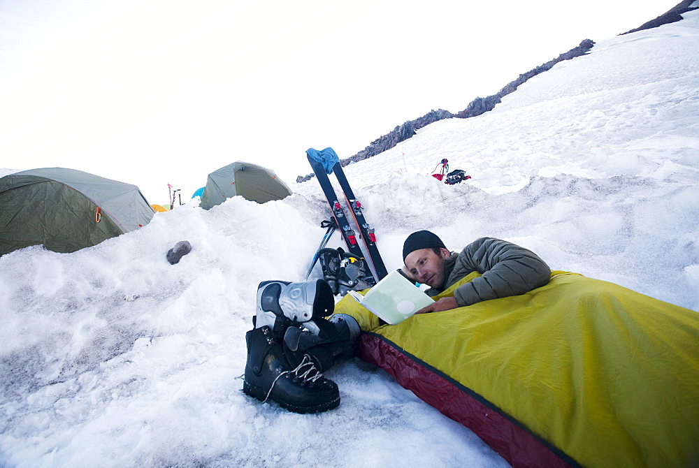 A young climber lies in his bivy sack and reads a book while the sun sets at base camp on Mount Rainier, United States of America