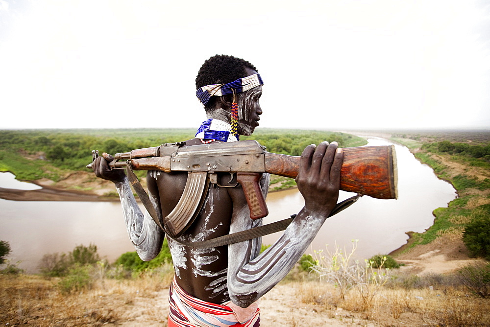 KARO VILLAGE, OMO VALLEY, ETHIOPIA. A young man holds his kalashnikov rifle while overlooking the Omo river in the remote Omo Valley of Ethiopia. (released: MHP_095_MR_RedKere), Ethiopia