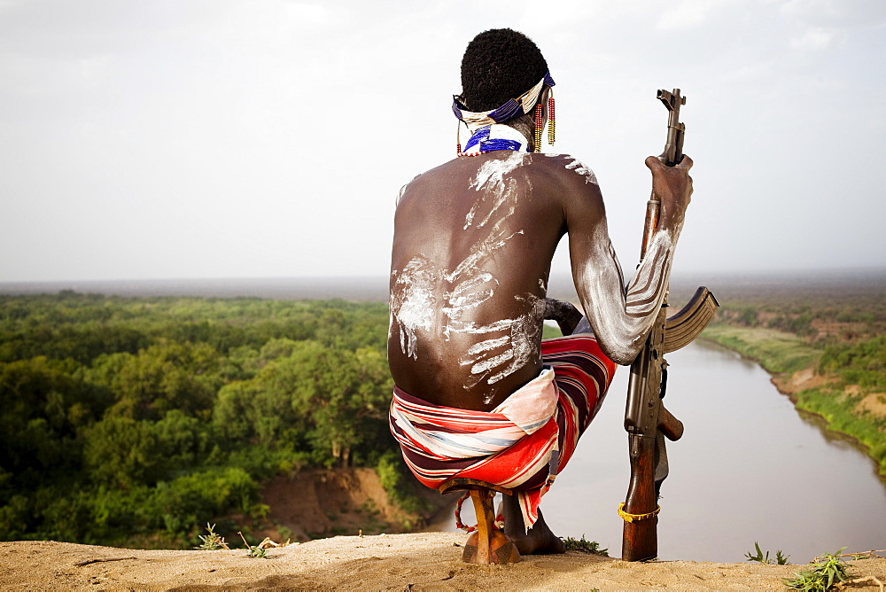 KARO VILLAGE, OMO VALLEY, ETHIOPIA. A young man holds his kalashnikov rifle while overlooking the Omo river in the remote Omo Valley of Ethiopia. (released: MHP_095_MR_RedKere), Ethiopia