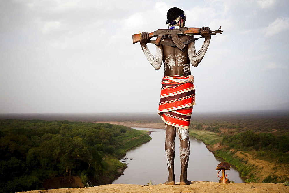 KARO VILLAGE, OMO VALLEY, ETHIOPIA. A young man holds his kalashnikov rifle while overlooking the Omo river in the remote Omo Valley of Ethiopia. (released: MHP_095_MR_RedKere), Ethiopia