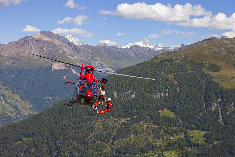 A mountain rescue emergency physician is hanging below a helicopter high above the ground. A injured hiker is being winched up. The Swiss Alps in the background.