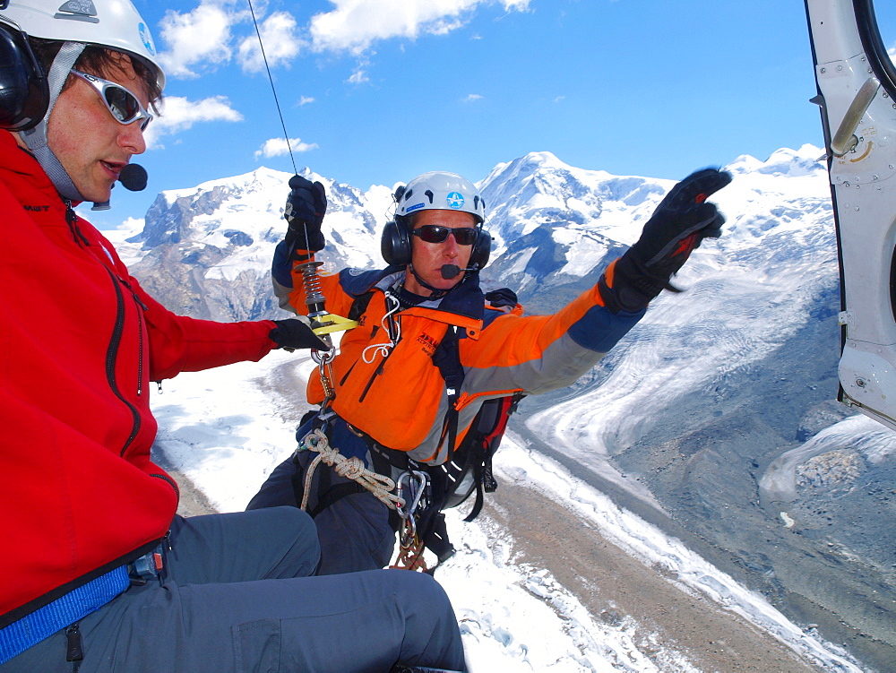 A mountain rescue technician is exiting a helicopter high above the ground. A winchman is ready to lower him down to the glacier where a climber is injured in a fall.