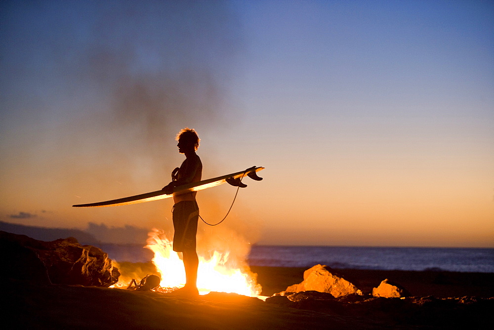 A surfer standing by a campfire on the beach in Hawaii.