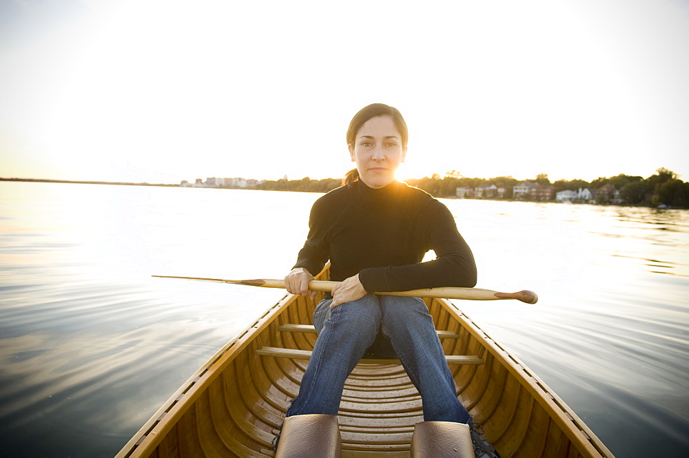 Mature Woman Holds Paddle On Lap In a Wood Canvas Canoe on a calm lake While Looking At Camera With Sun Setting In Background