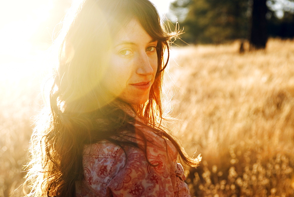 A backlit, young woman smiles and poses for a portrait in a large, grassy field near Flagstaff, Arizona.