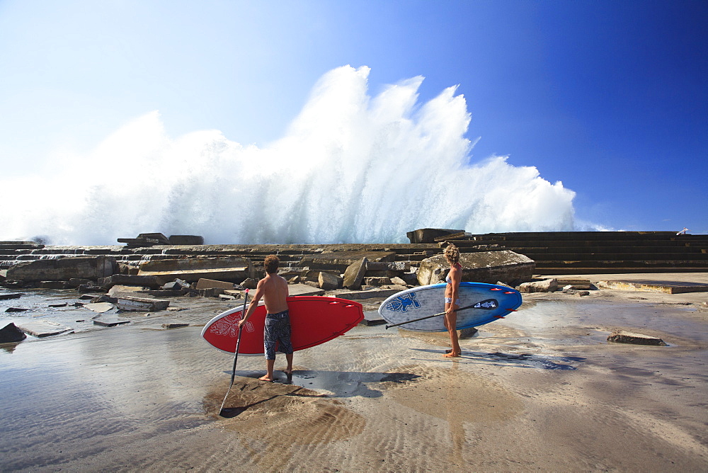 Pro SUP athletes Iballa Ruano and Vilayta watch the waves breaking against a pier in the Canary Islands.