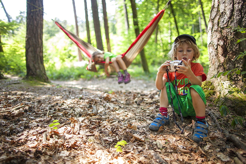 A 5 years old girl in an hammock, with her brother playing with a camera. In a forest, France.