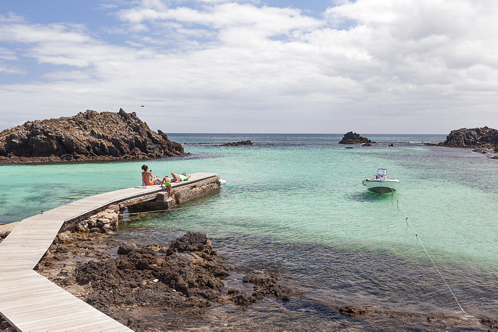 Family relaxing on a wooden jetty in Lobos Island (Fuerteventura) with a boat and clear calm blue ocean