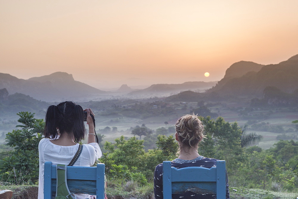 Two girls sitting on wooden chairs watching the sunrise above Vinales National Park from Los Acuaticos, the final point of an early morning trekking famous to watch the sun rising. Vinales, Cuba