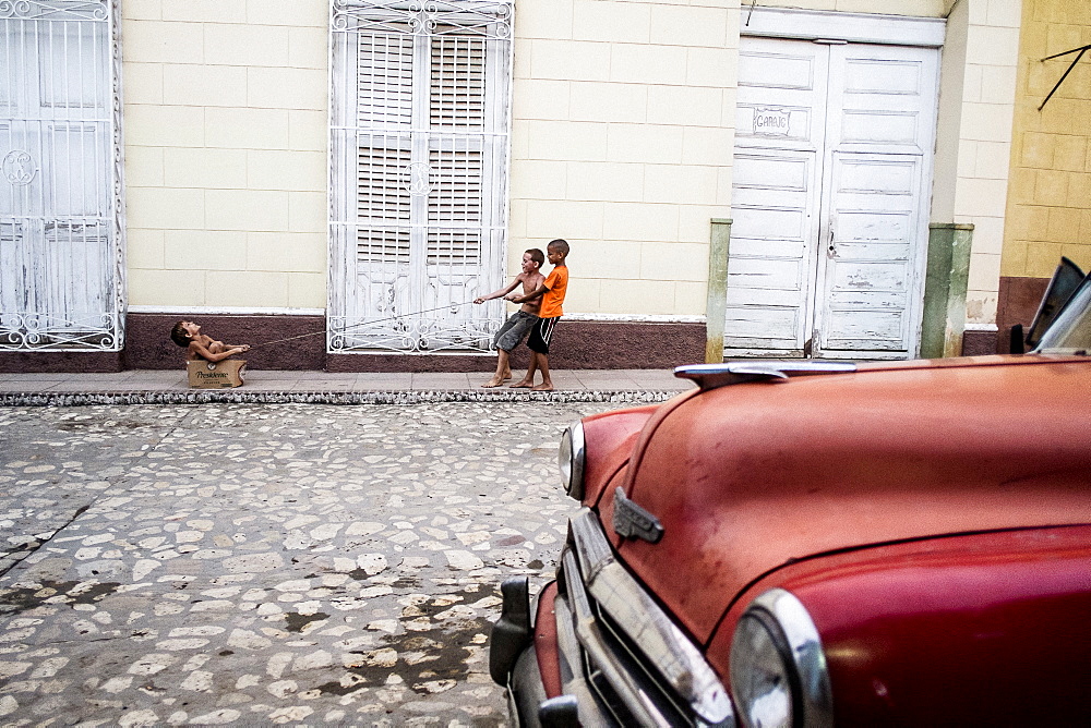 Three kids play on the street in front of a red classic car in Trinidad, Cuba