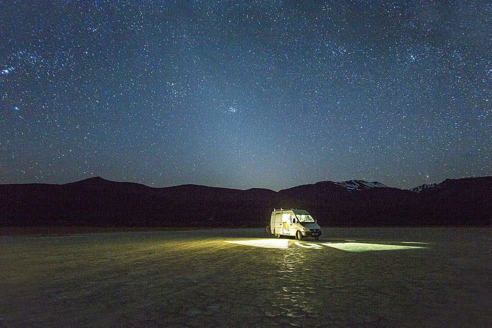 A white van is parked in a giant dried lake bed at night under a starry, Milky Way sky with what appears to be a halo of light around the van.